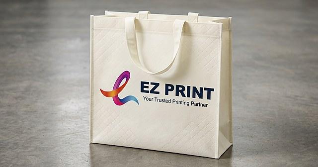 Photograph of a teal non-woven tote bag with matching fabric handles and a plain front print panel, standing on a table.